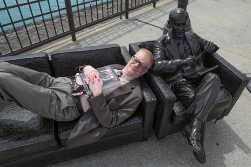 Author Walter Podrazik lies on the couch at the Bob Newhart sculpture on Navy Pier in Chicago with a copy of his book “Watching TV: American Television Season by Season,” on April 29, 2025. (Terrence Antonio James/Chicago Tribune)